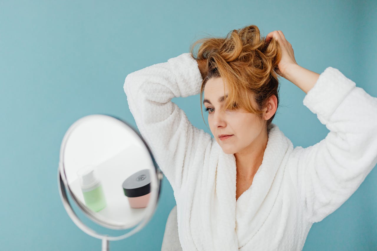 Woman in bathrobe looks in mirror while styling hair, in a modern indoor setting.
